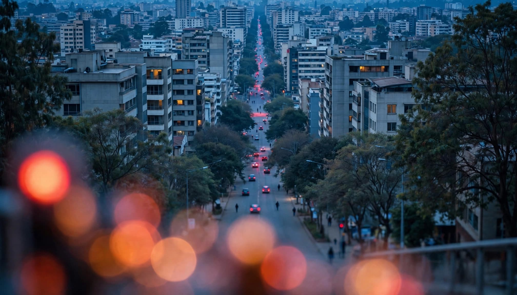 An elevated view of an urban street at dusk with buildings and bokeh lights in the foreground.